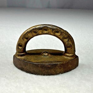 Puede incluir: Una pequeña plancha de metal de estilo antiguo con un acabado en bronce oscuro y desgastado. El objeto presenta un mango semicircular decorado con una serie de hendiduras circulares a lo largo del borde superior y una base plana y ovalada.