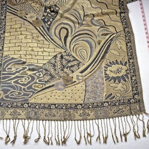 May include: A rectangular scarf featuring a gold and dark gray woven pattern with geometric shapes, floral motifs, and swirling lines. The scarf has a decorative border and fringe at the bottom edge, positioned next to a measuring tape showing 54 inches.