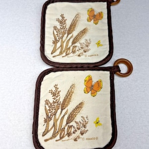 May include: Two square quilted pot holders with a cream-colored base, dark brown fabric trim, and a wooden hanging ring. The design features printed illustrations of brown wheat stalks, orange butterflies, and small yellow flowers with the text FRANCO.