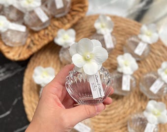 May include: Clear, seashell-shaped glass bottles, each topped with a white flower and a small tag. The bottles are arranged on a woven mat and in a wicker basket. The tag on the bottle in the foreground has text.