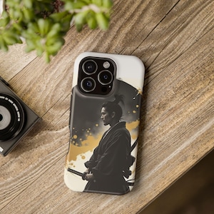 May include: A smartphone case featuring a black and white illustration of a samurai warrior. The design includes gold accents and a black background. The case is on a wooden surface.