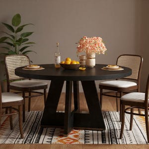 May include: A dark wood round dining table with four chairs. The chairs have woven backs and white cushions. The table is set with plates, a bowl of lemons, and a vase of pink flowers. A patterned rug is beneath the table.