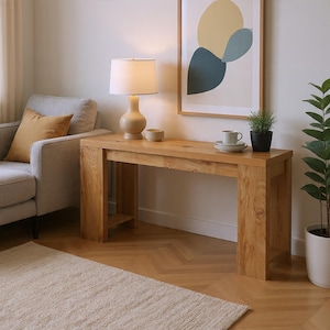 May include: A light oak wood console table with a lamp, small bowl, and a cup and saucer on top. A framed abstract art piece hangs on the wall behind the table. A gray sofa with a yellow pillow is to the left of the table. A potted plant is to the right.