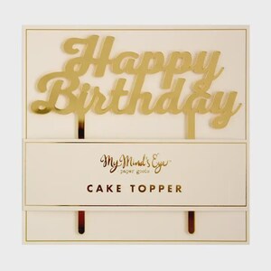 May include: A gold-colored cake topper featuring the words Happy Birthday in a cursive script font. The topper is attached to a rectangular card with the text My Mind's Eye paper goods and CAKE TOPPER printed in gold on the front surface.