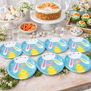 Puede incluir: Una mesa festiva de Pascua con una tarta de zanahoria, cupcakes y galletas decoradas. Platos azules con diseños de conejos y pollitos están dispuestos. Los platos tienen el texto "Happy Easter".