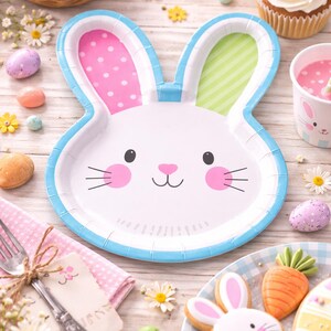 May include: An Easter table setting with a bunny-shaped plate, decorated with eggs, cupcakes, and cookies. The plate has pink polka dots and green stripes. The scene includes a pink drink and is set on a wooden surface.