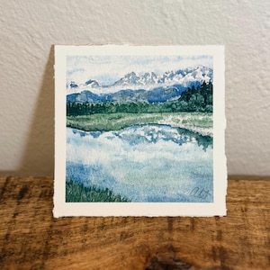 May include: A square watercolour painting on textured paper featuring a landscape with a calm blue lake in the foreground, a band of dark green forest in the middle ground, and snow-capped mountain peaks under a light blue sky. The artist's initials are visible.