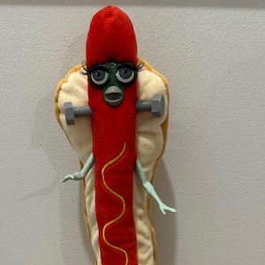 May include: A plush hot dog character with a red sausage, yellow mustard, and a bun. The character has a face with eyelashes and is holding bolts. The character has arms and legs. The text "goblin_milk_" is visible.