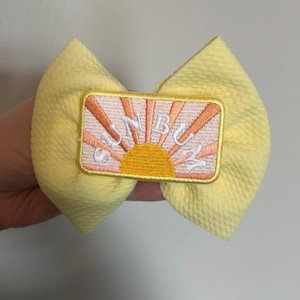 Pode incluir: Um laço de tecido amarelo com um patch bordado com um desenho de sol nascente. O patch tem o texto "SUNBUZZ" em letras brancas. O laço é de cor amarelo claro.