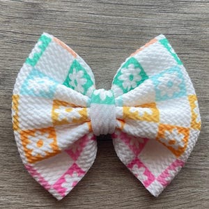 Può includere: Un fiocco per capelli fatto a mano con un colorato design patchwork su sfondo bianco. Il fiocco presenta un motivo ripetuto di margherite in quadrati arancioni, rosa, turchese e gialli. Il centro del fiocco è avvolto in tessuto bianco.