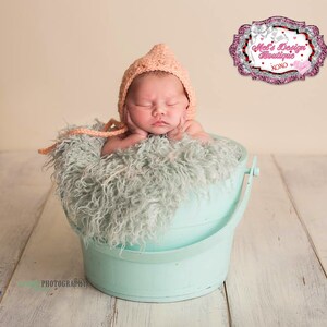 May include: A newborn baby wearing a peach-colored knitted bonnet, resting peacefully in a light blue bucket filled with fluffy, light green faux fur. The bucket sits on a white wooden surface. The image includes the text "Mel's Design Boutique XOXO".