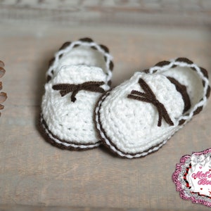 May include: A pair of handmade, white crochet baby shoes with brown accents. The shoes feature a brown trim, a decorative bow, and a scalloped edge. The shoes are designed to look like miniature loafers.