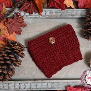 May include: A burgundy crochet cozy with a wooden button closure. The handmade item is displayed on a rustic wooden tray, surrounded by fall foliage, including leaves and pinecones. The cozy is likely for a coffee cup or similar item.