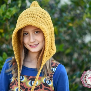 May include: A mustard yellow crochet hood with a pointed top, worn by a person. The hood has a textured, handmade appearance and is secured with ties. The person is wearing a blue shirt and a patterned dress. The background is blurred green foliage.