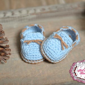 May include: A pair of baby booties, hand-crocheted in light blue yarn with brown trim and laces. The shoes are displayed on a wooden surface, with a pine cone in the background. The booties are a charming handmade item.