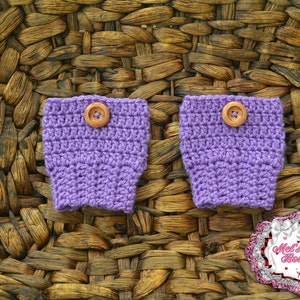 May include: Two lavender crochet wrist cuffs with wooden buttons. The cuffs are displayed on a woven brown background. The cuffs are handmade and feature a button closure. The image also includes a logo in the bottom right corner.