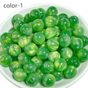 Op de afbeelding: Een verzameling doorschijnende groene kralen met een gemarmerd effect. De kralen zijn rond en lijken van glas of een vergelijkbaar materiaal te zijn gemaakt. De kralen zitten in een witte kom. De tekst "color-1" staat in de linkerbovenhoek.