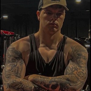 May include: A person wearing a black tank top and a brown and black trucker hat with the text "WORLD BASE". The person has tattoos on both arms.