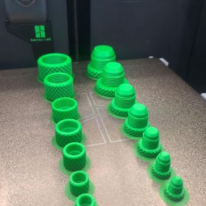 May include: A 3D-printed array of green plastic parts, including cylindrical and conical shapes with textured surfaces. The parts are arranged on a textured surface, likely a printer bed. The image showcases a variety of sizes and designs.