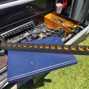 Könnte beinhalten: Ein schwarzer Steckschlüssel-Organizer mit gelben Innenräumen liegt auf einer blauen Oberfläche. Der Organizer ist mit kreisförmigen Öffnungen gefüllt. Im Hintergrund ist eine Werkzeugkiste mit verschiedenen Werkzeugen zu sehen, was auf einen Fokus auf Kfz- oder mechanische Werkzeuge hindeutet.