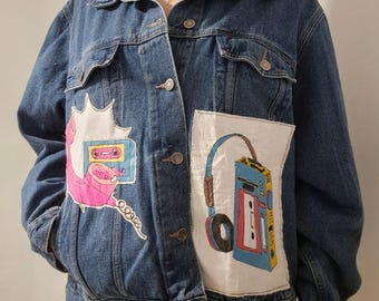 Peut inclure: Veste en jean bleue avec des motifs peints à la main de cassettes et de téléphones rétro. La veste a un col classique, des fermetures à boutons et des poches. L'œuvre d'art comprend des accents roses, bleus et jaunes, créant une esthétique vintage.