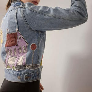 May include: Light blue denim jacket with a colorful, hand-painted design on the back. The artwork depicts a purple house with a red roof, a stop sign, and other buildings. The jacket has a classic button-up design.