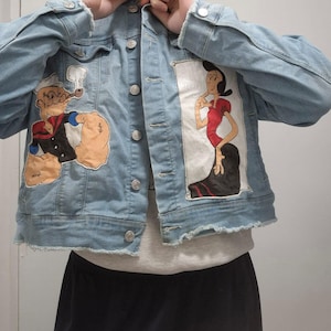 May include: Light blue denim jacket featuring cartoon characters Popeye and Olive Oyl. The jacket has a classic button-up design with silver buttons and frayed edges. The artwork is hand-painted on the front of the jacket.