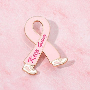 Puede incluir: Un pin de esmalte en forma de lazo rosa con las palabras "Keep Going" en escritura rosa. La cinta tiene zapatillas blancas adjuntas en la parte inferior. El pin tiene un borde dorado y está sobre un fondo rosa.
