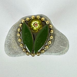 Könnte beinhalten: Ein dekorativer Stein mit floralem Design. Das Design umfasst zwei grüne Blattformen und eine kleine grüne Blume mit rotem Zentrum, umgeben von einer goldenen Perlenkette. Der Stein ist grau und hat ein natürliches, strukturiertes Aussehen.