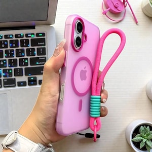 Może przedstawiać: Różowe etui na telefon z pasującym różowym paskiem. Etui ma okrągły wzór z logo Apple i pierścieniem magnetycznym. Pasek jest przymocowany z turkusowym akcentem. Etui na telefon jest pokazane obok laptopa.