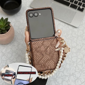 Op de afbeelding: Een bruine, getextureerde telefoonhoes met een slangenpatroon, ontworpen voor een Z Flip 7. De hoes heeft een groot venster en een parel- en gouden kettingriem met decoratieve bedels. De telefoon is open en toont het scherm.