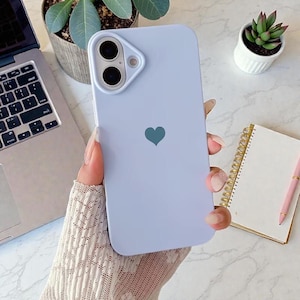 May include: A light purple phone case with a teal heart design. The case has a camera cutout and is held by a person. The background includes a laptop, a small succulent, and a notebook with a pink pen.