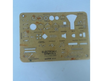May include: A vintage brass-colored stencil featuring various electronic symbols, including sockets, tubes, and capacitors. The stencil includes text that reads "ELECTRONIC SYMBOLS" and "I.E.C. STANDARDS".