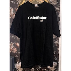 Puede incluir: Camiseta negra con el texto blanco "CodeWarrior" en el pecho. La camiseta está colgada en una percha de madera. El fondo es un papel pintado floral.