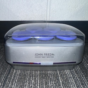 Op de afbeelding: Een zilverkleurige John Frieda haarroller-set met een doorzichtig plastic deksel, bevattende zes paarse rollers. De voorzijde van het apparaat toont de tekst John Frieda London Paris New York, een rood indicatielampje en een witte schakelaar.