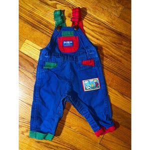 May include: Blue denim baby overalls with red and green accents. The overalls have a red pocket and green trim. A small patch with a cartoon scene is sewn on the leg. The brand name "OshKosh" is visible on the chest pocket.
