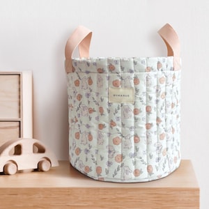 May include: A quilted storage basket with a floral and bunny pattern in soft pastel colors. The basket has peach-colored handles and a label that reads "BYMARUE". The basket is sitting on a wooden surface.