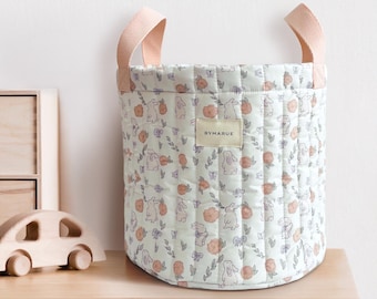 May include: A quilted storage basket with a floral and bunny pattern in soft pastel colors. The basket has peach-colored handles and a label that reads "BYMARUE". The basket is sitting on a wooden surface.