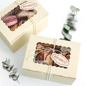 May include: Two beige bakery boxes with scalloped window cutouts, tied with string, and labeled "BON APPETIT." One box contains a pink macaron and other pastries. The other box holds cinnamon rolls. Eucalyptus sprigs are in the background.