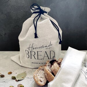 May include: A beige linen bread bag with a black drawstring closure. The bag is printed with the words "Homemade BREAD" in a stylish font. Sliced bread and a white cloth are in front of the bag.