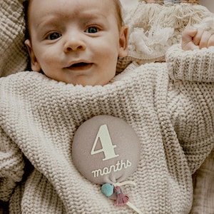 a photo of baby girl smiling with balloon shaped stuffed monthly milestone sign in dusty pink with blue pom pom with ivory felt that says "4 months". unique baby shower gift for new mom, parents. great alternative to traditional milestone blanket, paper card signs, wooden discs or blocks.
