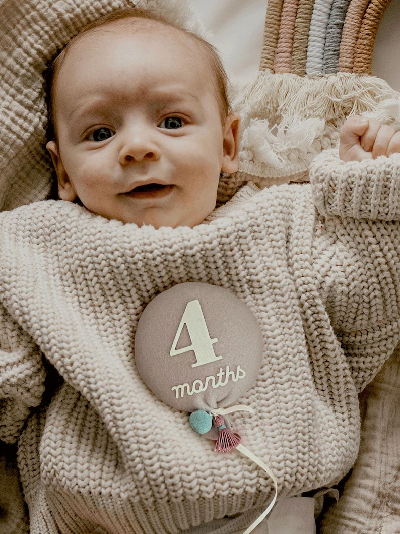 a photo of baby girl smiling with balloon shaped stuffed monthly milestone sign in dusty pink with blue pom pom with ivory felt that says "4 months". unique baby shower gift for new mom, parents. great alternative to traditional milestone blanket, paper card signs, wooden discs or blocks.