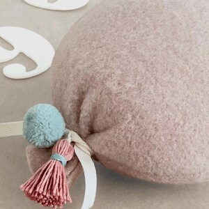 May include: A dusty rose felt balloon decoration featuring a soft, textured surface. The base of the balloon is tied with a white ribbon and adorned with a small, light blue pom-pom and a matching dusty rose tassel with a light blue thread accent.
