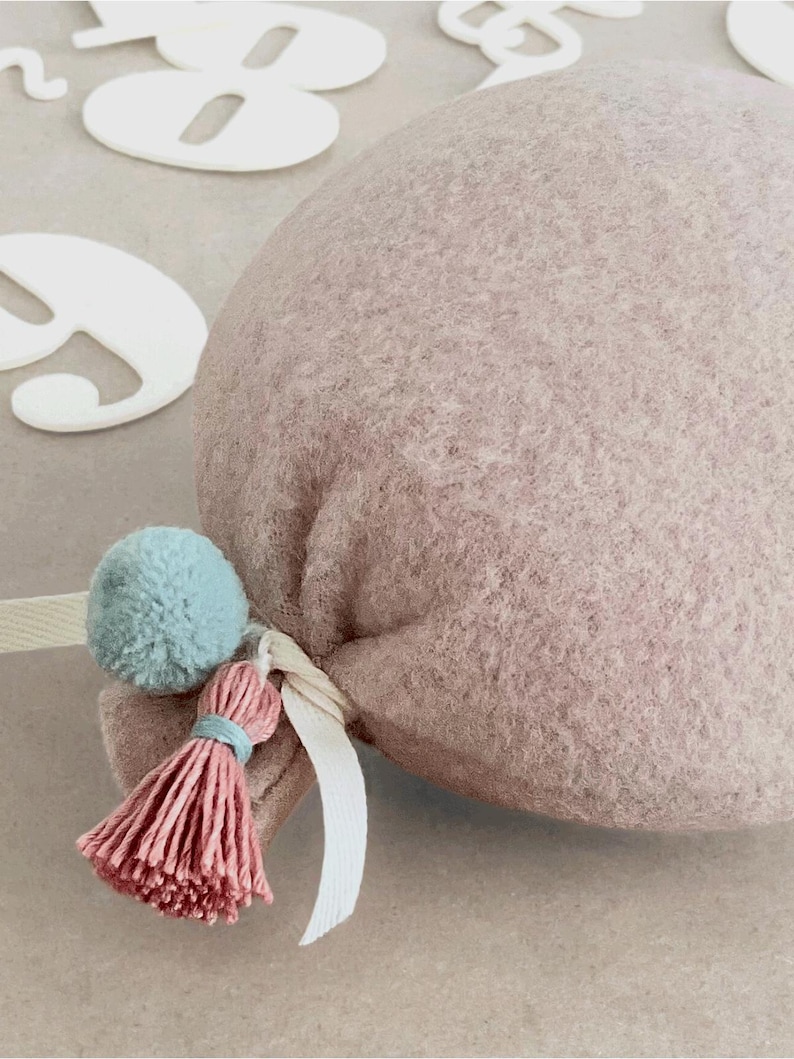May include: A dusty rose felt balloon decoration featuring a soft, textured surface. The base of the balloon is tied with a white ribbon and adorned with a small, light blue pom-pom and a matching dusty rose tassel with a light blue thread accent.