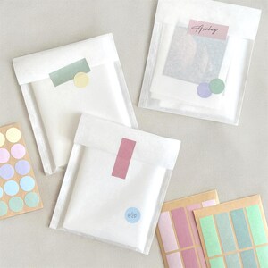 May include: Three white paper pouches with colorful stickers and labels. One pouch has a pink label and a blue circle sticker with "4/20". Another has a green label and a yellow circle sticker. The third pouch has a pink label with the name "Ashley".