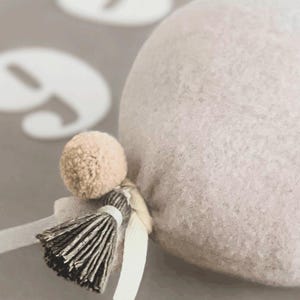 May include: A round, light beige felted object featuring a small, matching beige pom-pom and a dark grey, multi-strand tassel attached with a light-colored ribbon. The texture of the felted material is visible, and the object is rounded in shape.