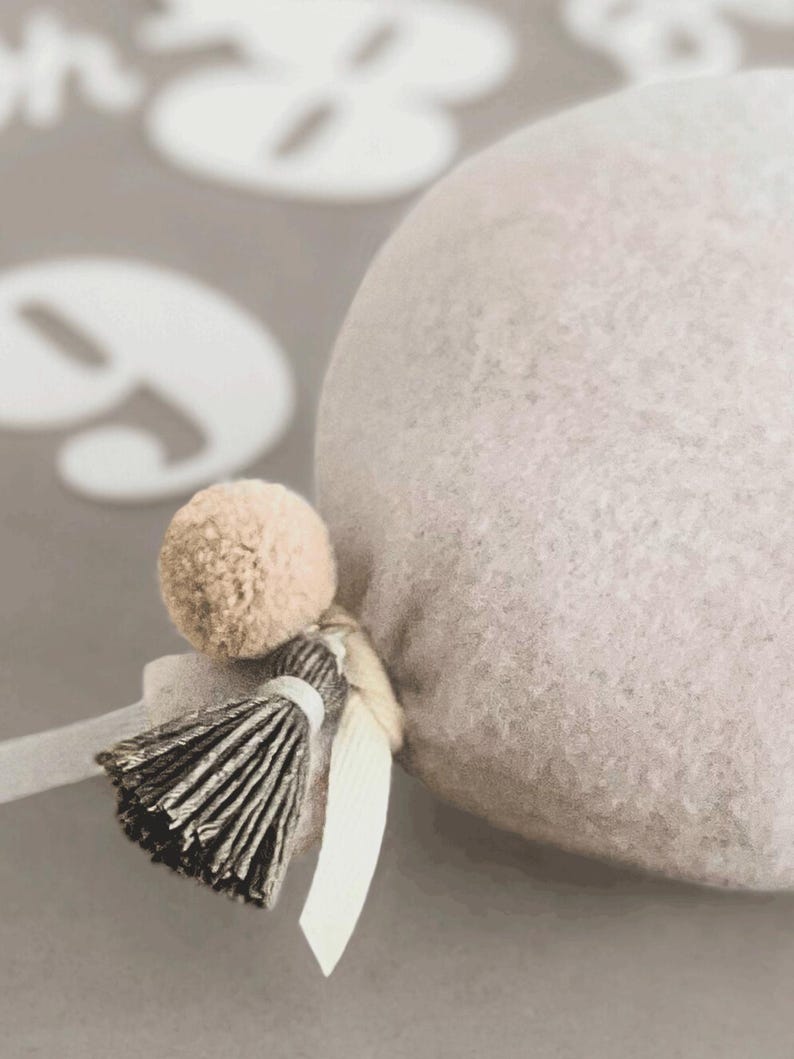 May include: A round, light beige felted object featuring a small, matching beige pom-pom and a dark grey, multi-strand tassel attached with a light-colored ribbon. The texture of the felted material is visible, and the object is rounded in shape.