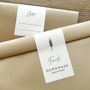 May include: Two tan paper-wrapped packages featuring white rectangular labels with serrated edges. One label displays a wheat stalk illustration, the text "Fresh" and "HOMEMADE BAKED GOODS". The other label shows the text "Enjoy!" and two blank lines.