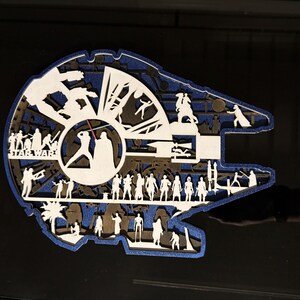 Könnte beinhalten: Ein mehrschichtiges Wandkunstobjekt aus Holz in Form des Millennium Falcon Raumschiffs. Das Design zeigt filigrane, weiß lasergeschnittene Silhouetten von Star Wars Charakteren und Szenen vor einem dunkelblauen Glitzerhintergrund und schwarzen Ebenen.