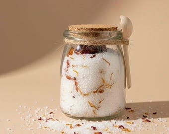 May include: A clear glass jar filled with white bath salts and dried flower petals, topped with a cork lid and a small wooden spoon. The jar is tied with twine and sits on a beige surface, with scattered salt crystals.